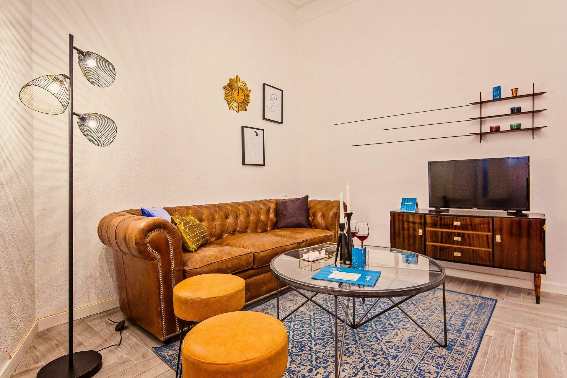 Una sala de estar con un sofá de cuero marrón, dos otomanas amarillas, una mesa de centro de vidrio, una lámpara de pie, un televisor y un mueble de madera.