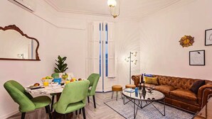 Una sala de estar con un sofá de cuero marrón, sillas verdes, una mesa de centro y una mesa de comedor con fruta y un libro.