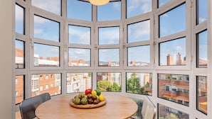 Ein Zimmer mit einem großen Erkerfenster, einem runden Holztisch mit einer Obst- und Gemüseanordnung sowie einer modernen Leuchte.