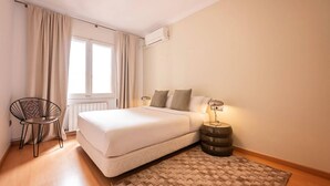 A bedroom with a bed, a chair, a bedside table with a lamp, and a window with curtains.