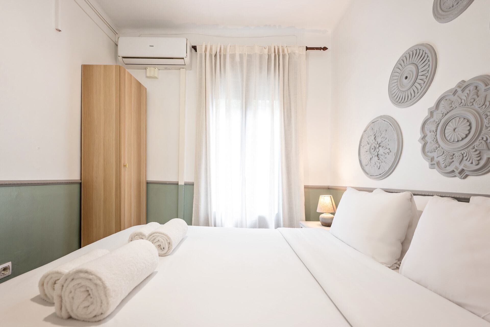 A hotel room with a bed, a wooden wardrobe, a window with curtains, and decorative wall plates.