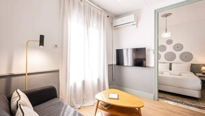 A modern hotel room with a sofa, a wooden table, a TV mounted on the wall, and a bed with patterned curtains.
