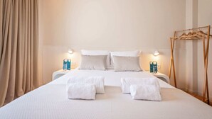 Une chambre d’hôtel avec un grand lit, deux tables de chevet, un porte-manteau en bois et des rideaux de couleur beige.