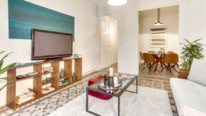 A modern living room with a flat-screen TV, a glass coffee table, and a dining area with yellow chairs.