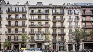 A row of multi-story buildings with balconies, parked cars, and motorcycles on the street.