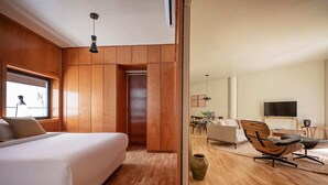 A modern bedroom with a large bed, wooden paneling, a window with blinds, a desk, a chair, and a television. - Sweett | Bogatell Beach (Barcelona)