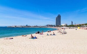 Una playa con gente tomando el sol y nadando, un barco grande al fondo y un edificio alto.