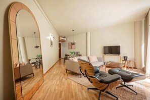 Un salon spacieux avec un grand miroir, un canapé, deux fauteuils, un téléviseur et un coin repas comprenant une table et des chaises.