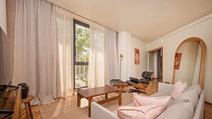 A living room with a sofa, a coffee table, a chair, and a lamp. There is a window with curtains and a view of trees outside. - Sweett | Bogatell Beach (Barcelona)