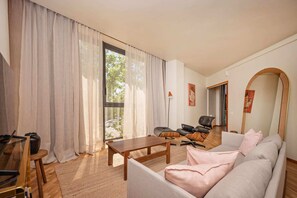 Un salon avec un canapé, une table basse, un fauteuil et une lampe. Il y a une fenêtre avec des rideaux et une vue sur les arbres à l’extérieur.