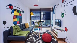 Een moderne woonkamer met een bank, een bijzettafeltje en een rode poef. Er is een raam met uitzicht op de stad.