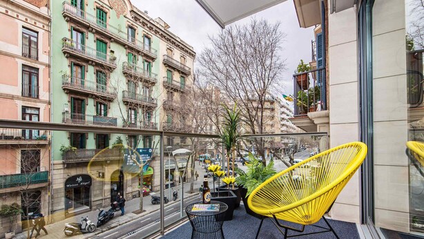 Een balkon met een gele stoel, een kleine tafel en een glazen hekwerk, met uitzicht op een straat met gebouwen en geparkeerde scooters.