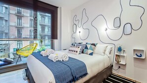 Een moderne slaapkamer met een bed, kussens, een blauw-wit geborreld deken, een gele stoel en een uitzicht op gebouwen door een raam.