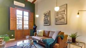 Una sala de estar acogedora con un sofá de madera, una mesa de centro de vidrio y un tapete colorido. Se ve un ventanal con persianas de madera, un maniquí y cuadros en las paredes.