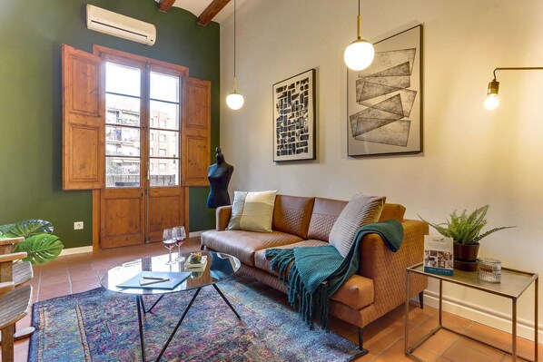 A cozy living room with a wooden sofa, a glass coffee table, and a colorful rug. There is a large window with wooden shutters, a mannequin, and framed artwork on the walls.