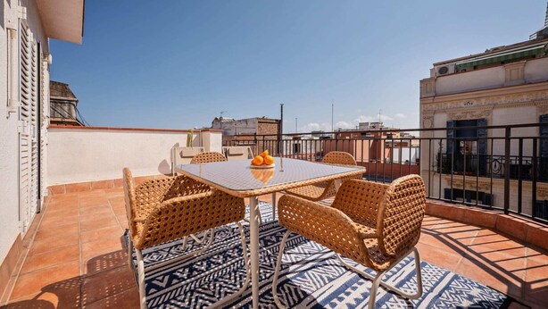 Una terraza en la azotea con una mesa y cuatro sillas de mimbre, un tapete estampado y una planta de cactus.
