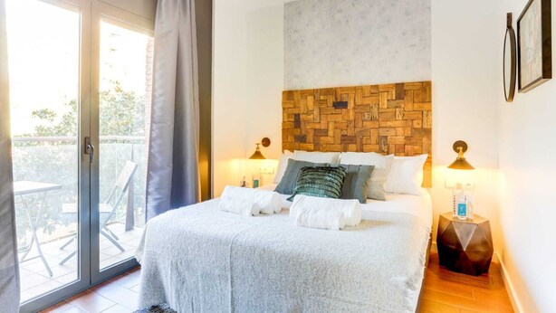 A bedroom with a large bed, a wooden headboard, two bedside lamps, and a view through a sliding glass door to an outdoor area. - Sweett | Raul - Two Bedroom Apartment, Sleeps 4 (Barcelona)