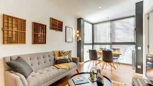 A modern living room with a grey sofa, a round coffee table, and a dining area with a table and chairs. There are large windows with blinds, a framed picture on the wall, and a radiator on the floor. - Sweett | Raul - Two Bedroom Apartment, Sleeps 4 (Barcelona)