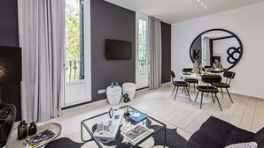 A modern living room with a dining area, large windows, a flat-screen TV, and a circular wall decoration.