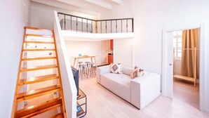 A modern loft apartment with a staircase leading to a mezzanine, a living area with a sofa and a dining table, and a kitchen in the background. - Sweett | Ethnic Eixample (Barcelona)