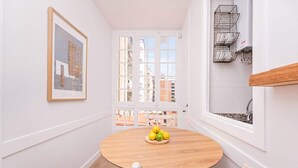 Una habitación pequeña y luminosa, con una mesa redonda de madera y dos sillas, un cuadro en la pared, una ventana con vistas a edificios, y una zona de cocina con calentador de agua.