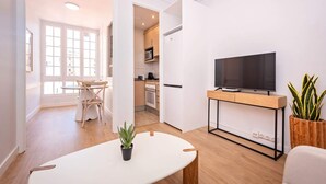 Una sala de estar moderna con un televisor de pantalla plana sobre un soporte de madera, una zona de comedor con una mesa y sillas, y una cocina al fondo.
