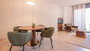 A modern living room with a round wooden table, green upholstered chairs, a TV stand, and a large window with curtains. - Sweett | Cosy Ciutadella (Barcelona)