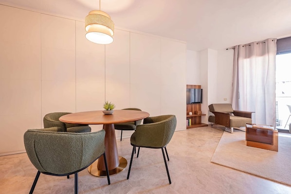 Un salon moderne avec une table ronde en bois, des fauteuils à revêtement vert, un meuble pour la télévision et une grande fenêtre avec des rideaux.