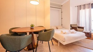 A hotel room with a round wooden table, two green chairs, a bed with white linens, a small potted plant, and a hanging light fixture. - Sweett | Cosy Ciutadella (Barcelona)