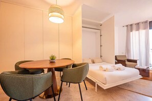 A hotel room with a round wooden table, two green chairs, a bed with white linens, a small potted plant, and a hanging light fixture.