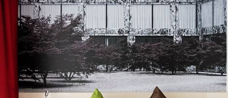A hotel room with a bed, two pillows, a nightstand, a red curtain, and a large wall art depicting trees and a building.