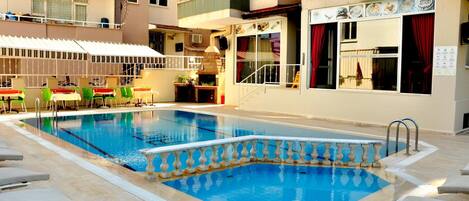 A swimming pool with lounge chairs, a covered seating area, and a building with balconies and windows.