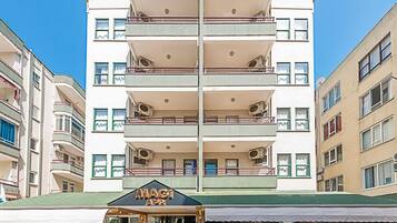 A multi-story hotel with a central entrance, balconies on each floor, and outdoor seating with potted plants.