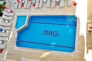 Een zwembad met 'MAGI' op het wateroppervlak, omringd door ligbedden en lounge fauteuils.