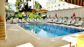 A swimming pool with lounge chairs and umbrellas, surrounded by a building with balconies and greenery.