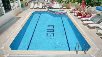 Ein Schwimmbecken mit der Aufschrift 'MAGI' in der Mitte, umgeben von Liegestühlen und einer Poolanlage.