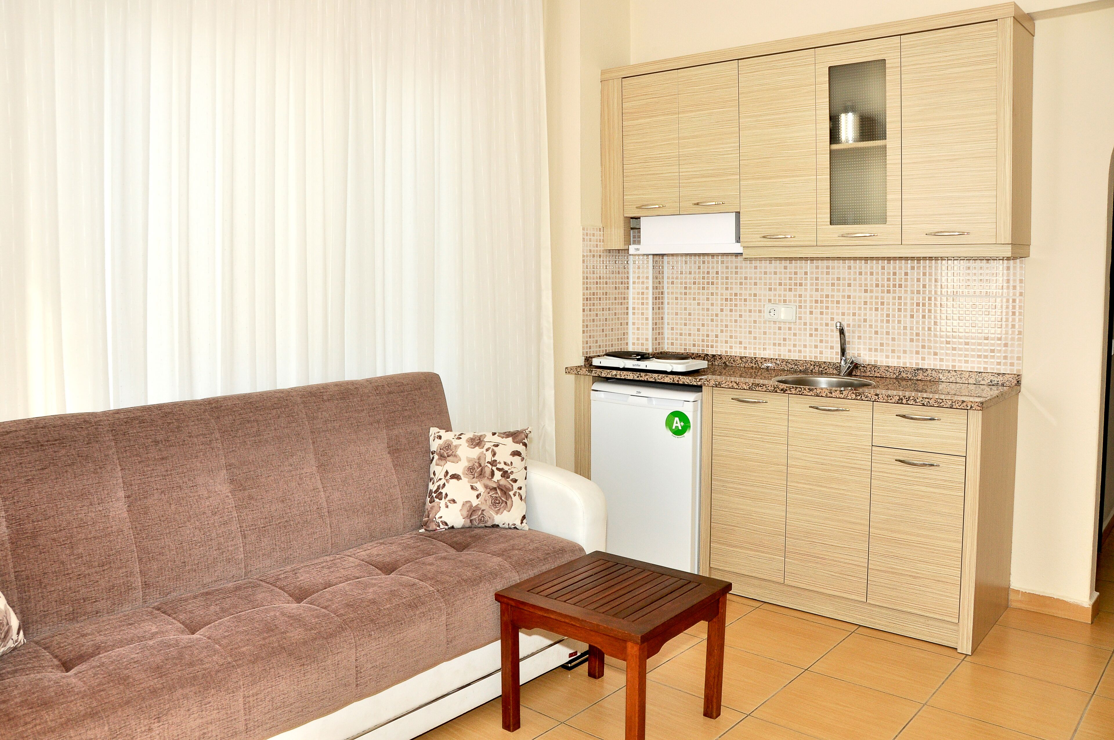 Un espacio habitable pequeño con sofá, una mesita de madera, una kitchenette con refrigerador y una ventana con persianas.