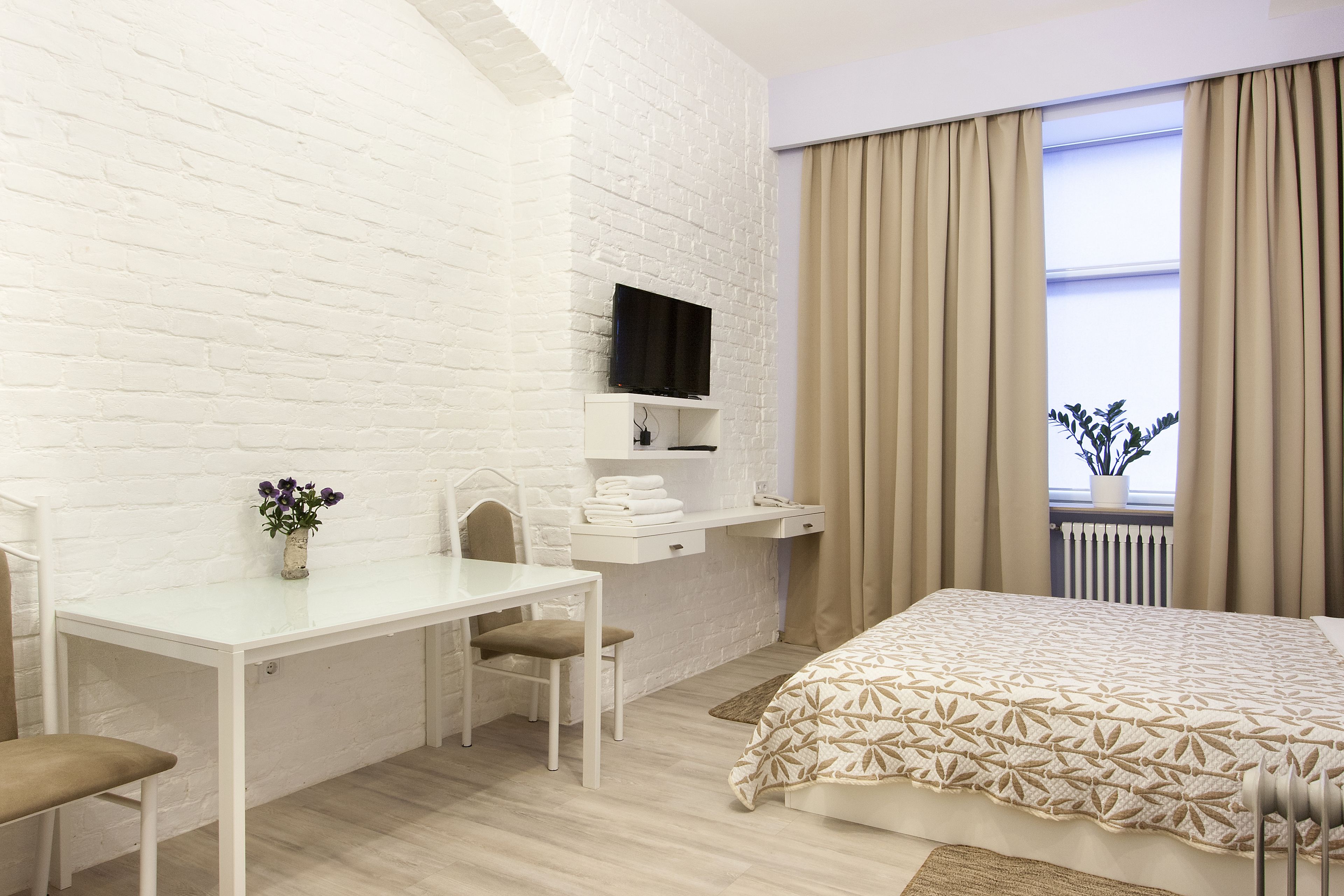 Une chambre d’hôtel moderne avec un mur en briques blanches, un téléviseur à écran plat fixé au-dessus d’un bureau, un lit avec une couette à motifs et une fenêtre ornée de rideaux beiges.