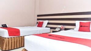 Habitación de hotel con dos camas individuales, cada una con un almohadón rojo y una colcha blanca. Tiene un cabecero de madera, una mesita de noche con un teléfono y una manta roja en el suelo.