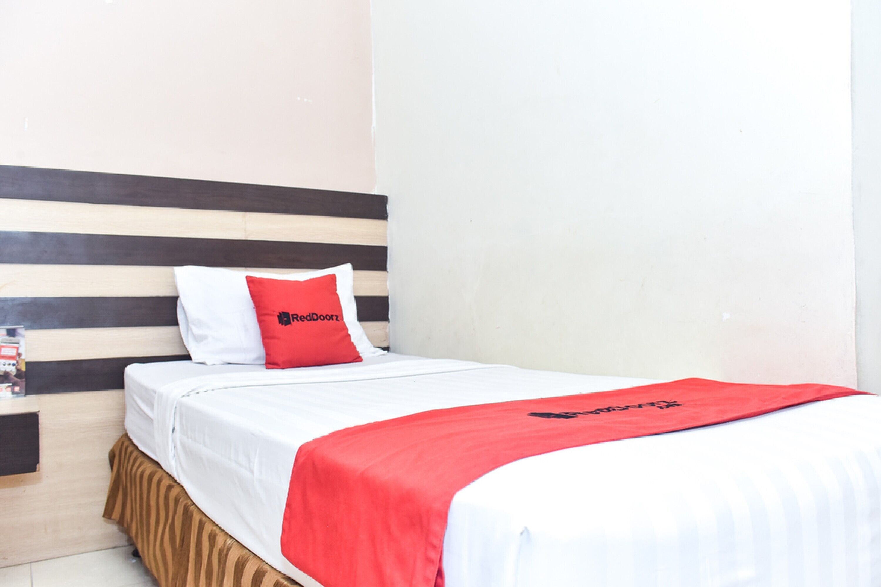 A hotel room with a bed, a red and white pillow, a red blanket, and a wooden headboard.
