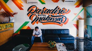 Un vestíbulo de hotel con un llamativo diseño mural geométrico, un sofá, una mesa de centro con un jarrón de flores y un periódico.