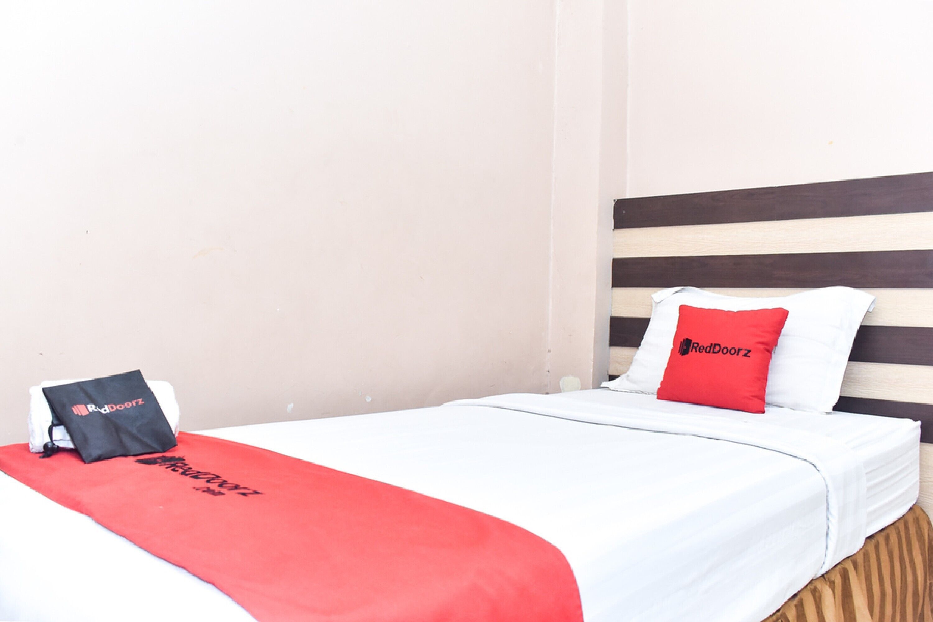 A hotel room with a bed, a red and white bedspread, a black bag, and a pillow with the RedDoorz logo.