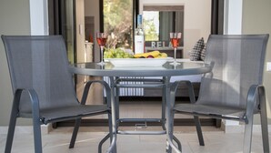 A round glass table with a white plate of fruit and a bottle of wine, flanked by two grey mesh chairs. - Gmare (Polygyros)