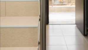 A modern staircase with beige steps and a metal handrail, leading to a bright interior space with a glass door and a visible exit sign. - Gmare (Polygyros)