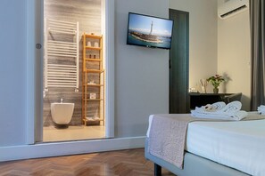A modern hotel room with a large bed, a television mounted on the wall, a wooden shelf with toiletries, and a view of a lighthouse through the window. - Re Vittorio Deluxe (Messina)