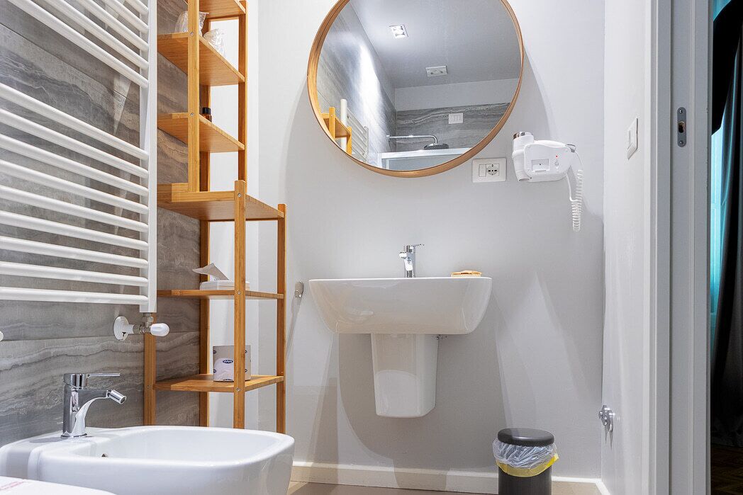 A bathroom with a white sink, a mirror, a wooden shelf unit, and a trash bin.