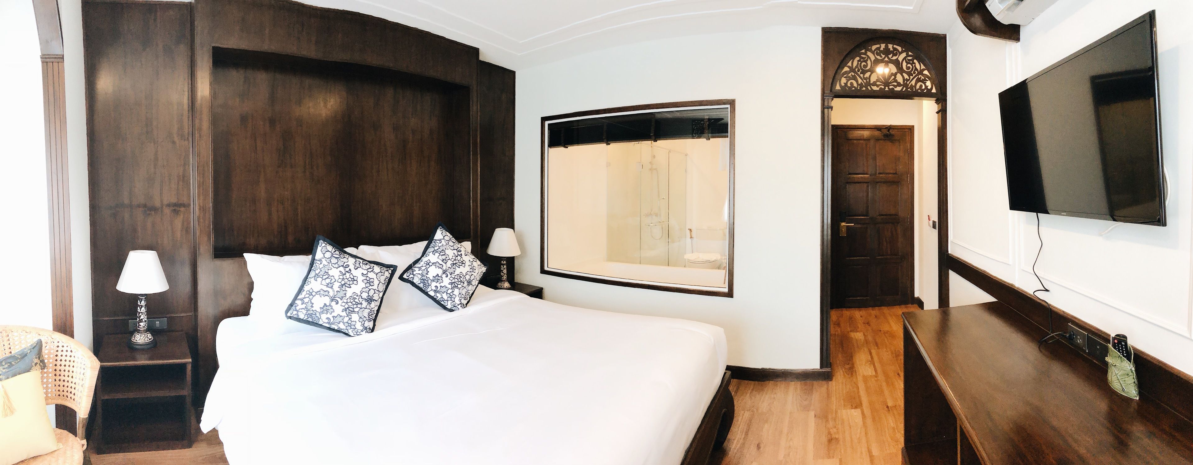 A hotel room with a large bed, a TV, a wooden floor, and a bathroom visible through a glass door.