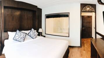 A hotel room with a large bed, a TV, a wooden floor, and a bathroom visible through a glass door.