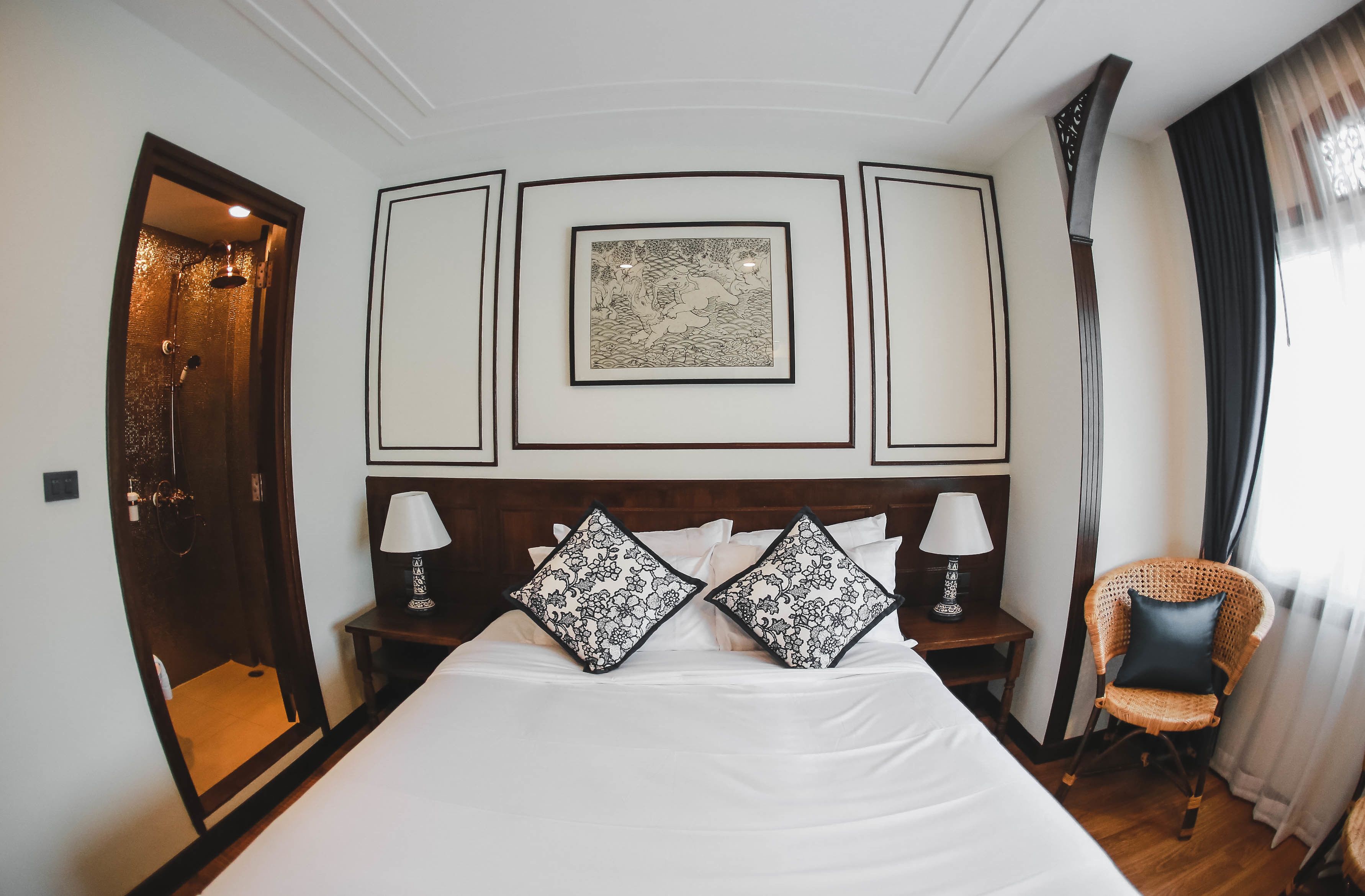 Una habitación de hotel con una cama grande, dos mesitas de noche con lámparas, un panel decorativo en la pared, una silla de mimbre y una zona de ducha.