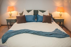 Cama bem arrumada com um cobertor azul de tricô, duas mesas de cabeceira com luminárias e cabeceira com várias almofadas.